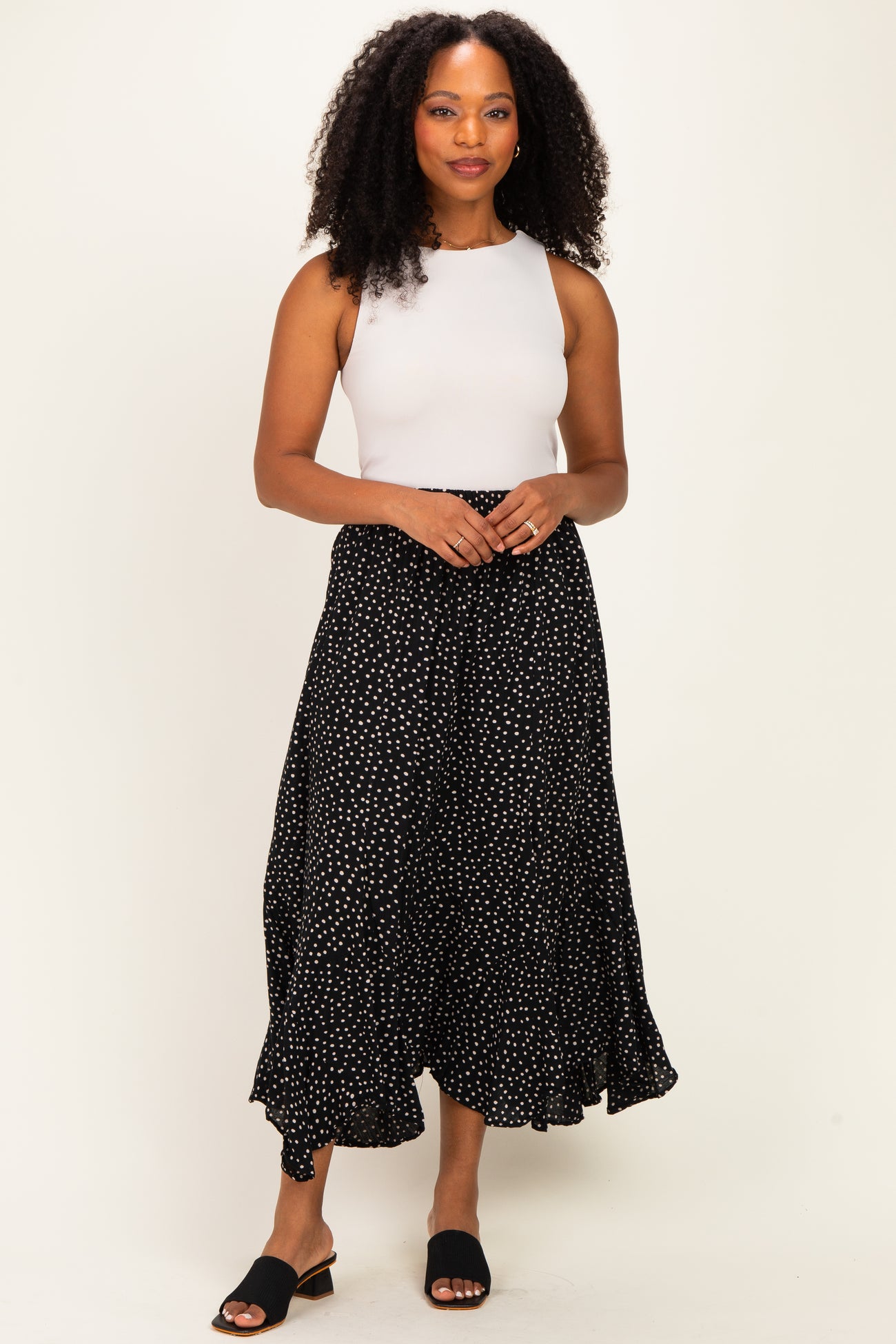 woman wearing a white sleeveless top and a black polka dot maxi skirt, standing confidently with hands clasped