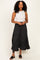 woman wearing a white sleeveless top and a black polka dot maxi skirt, standing confidently with hands clasped