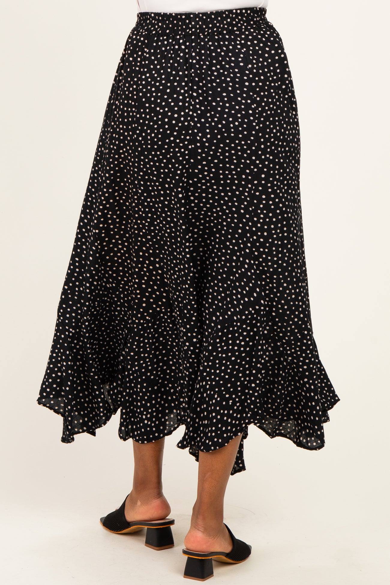 woman wearing a black polka dot midi skirt with a ruffled hem and back elastic waistband, paired with black heeled mules