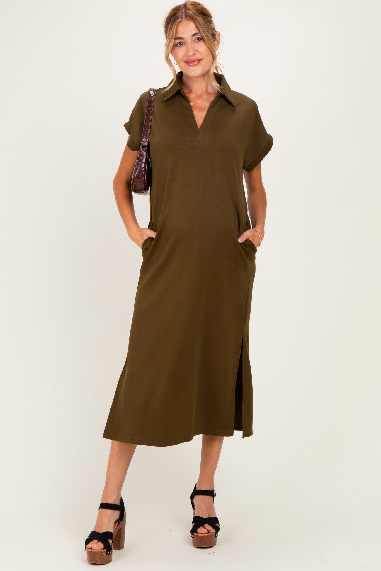 woman wearing an olive green short sleeve maternity dress with a collar, side slits, and pockets, holding a handbag
