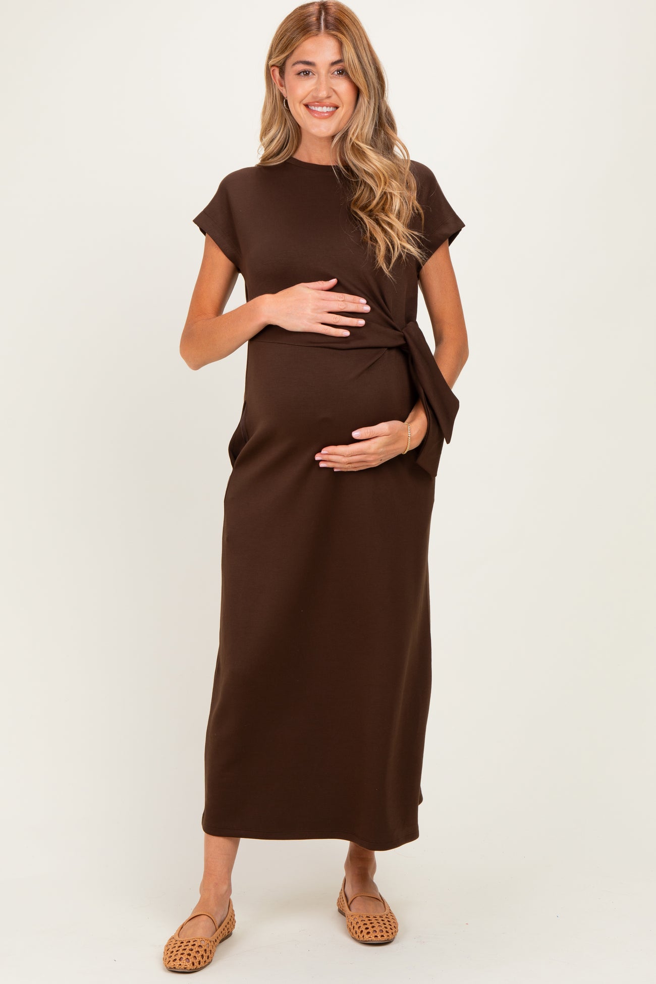 woman wearing a brown maternity dress with a side tie, holding her baby bump, standing against a neutral background
