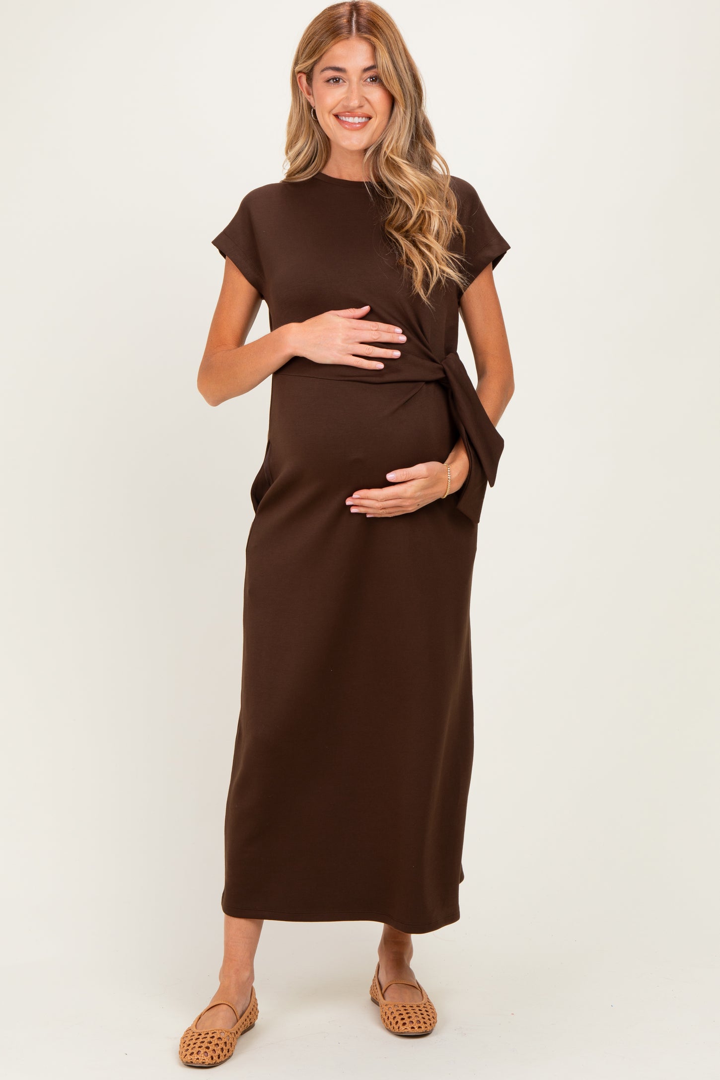 woman wearing a brown maternity dress with a side tie, holding her baby bump, standing against a neutral background