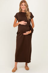 woman wearing a brown maternity dress with a side tie, holding her baby bump, standing against a neutral background