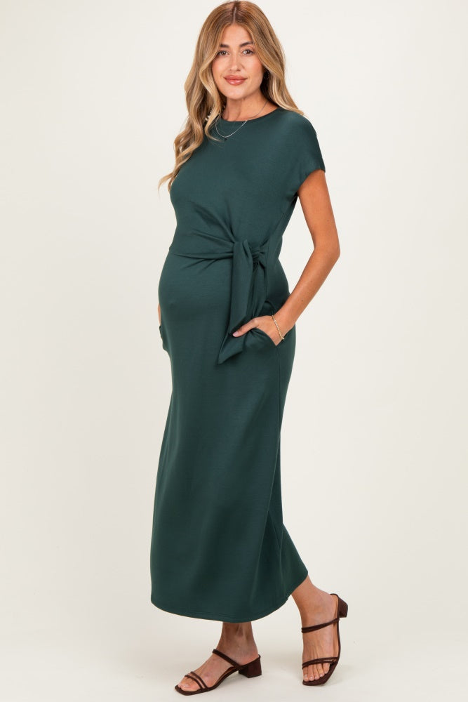 woman wearing a dark green maternity maxi dress with a tie waist and pockets, showing her baby bump