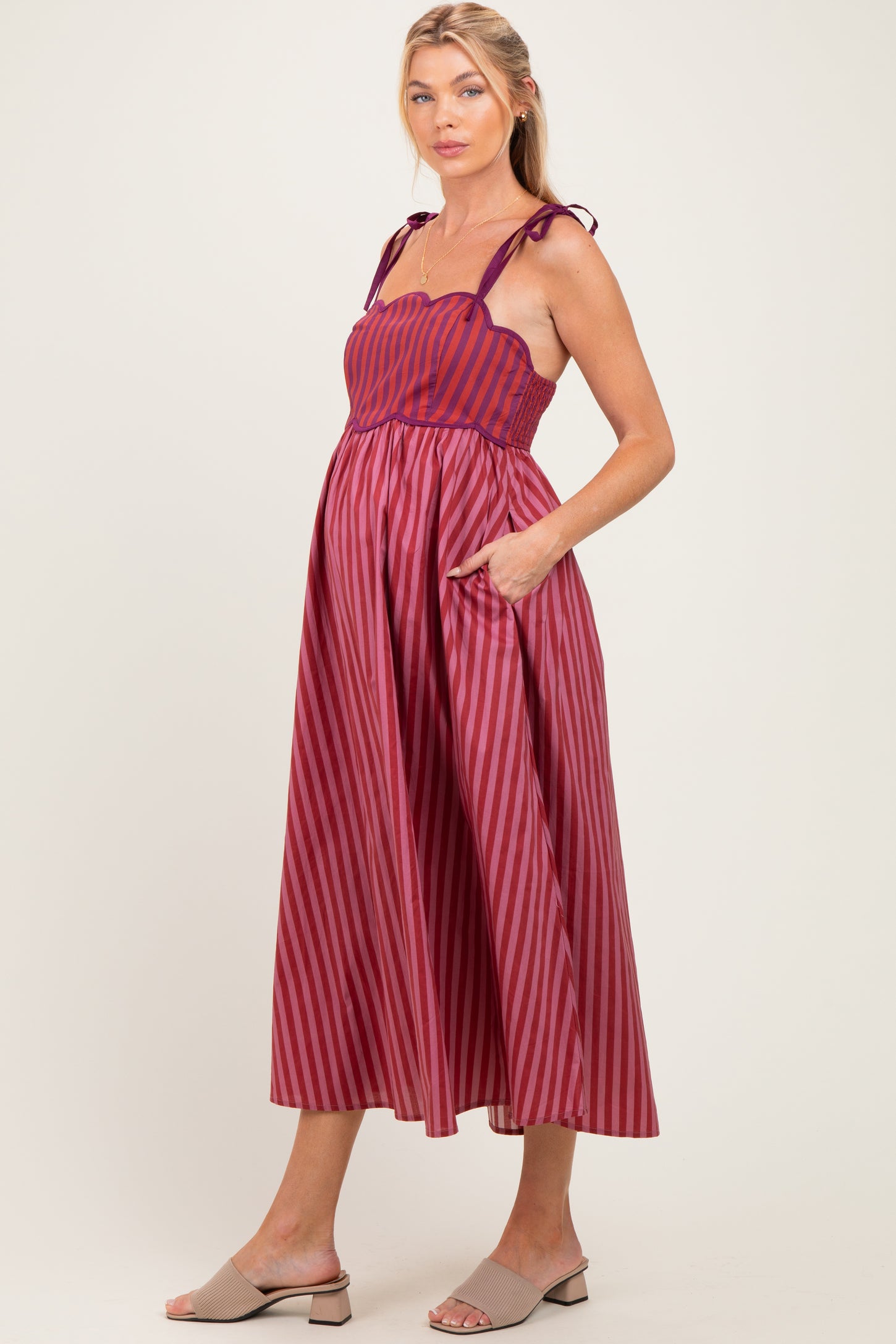 woman wearing a burgundy striped maternity dress with adjustable straps and pockets, showcasing a baby bump