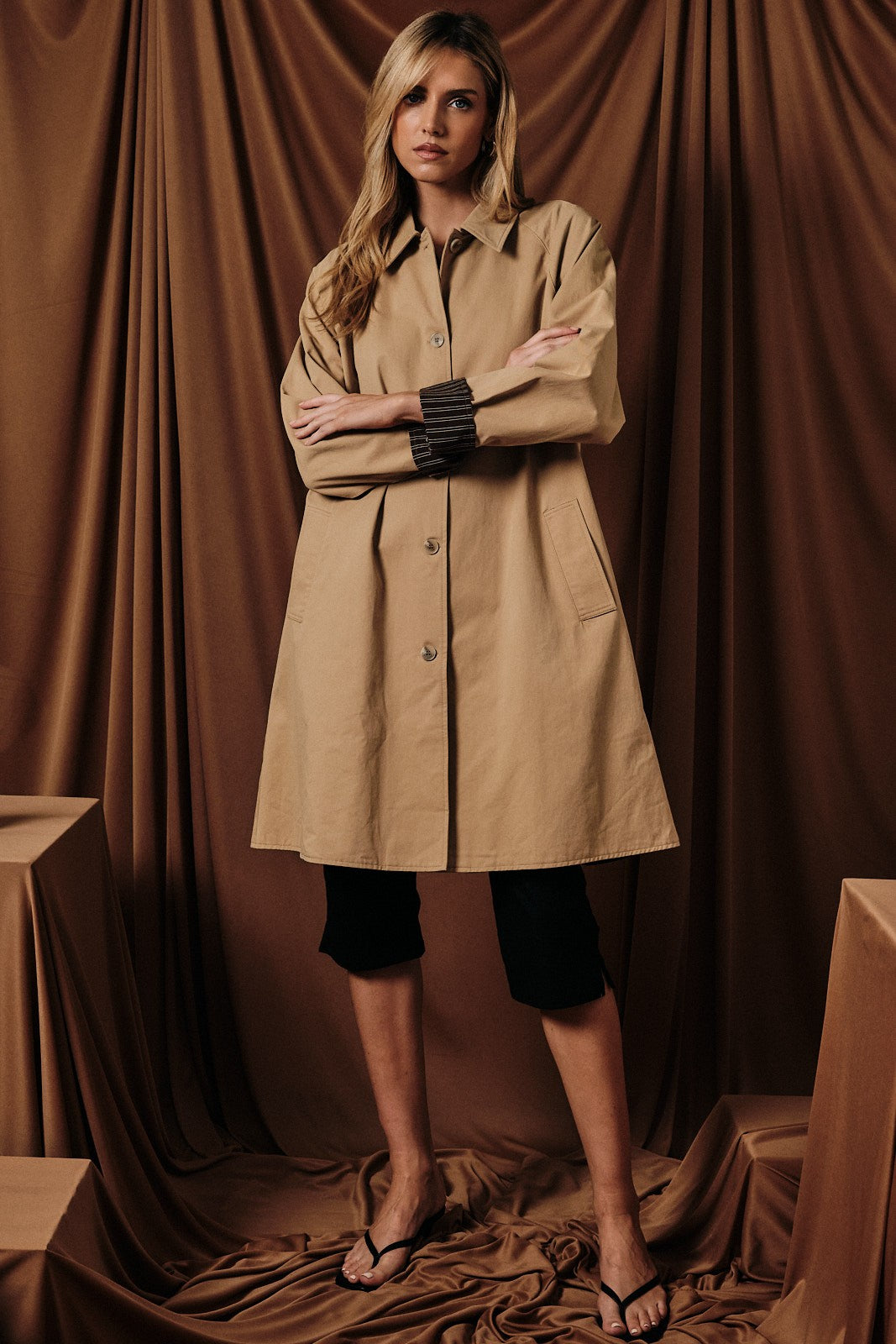 woman wearing a beige oversized coat over black cycling shorts, standing against a brown backdrop