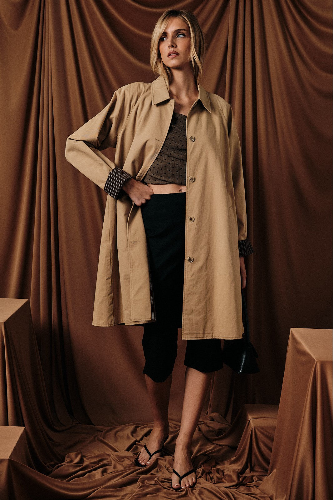 woman wearing a tan oversized trench coat over a brown cropped top and black capri pants, posing against a draped background