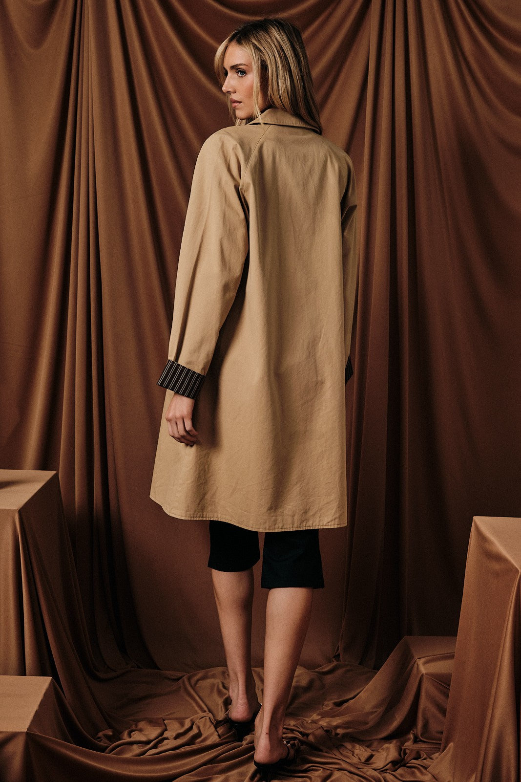 woman wearing a tan long sleeve coat with striped cuffs, standing with her back to the camera against a brown backdrop
