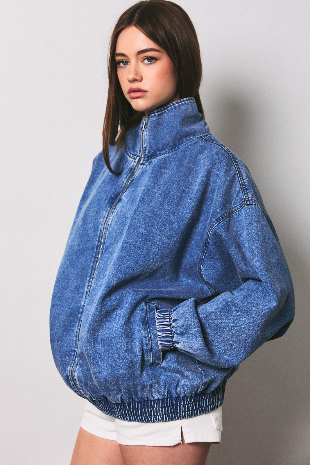 woman wearing an oversized blue denim zip-up jacket with a high collar and elasticized cuffs, paired with white shorts