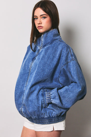 woman wearing an oversized blue denim zip-up jacket with a high collar and elasticized cuffs, paired with white shorts