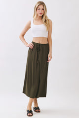 woman wearing an olive green maxi skirt with an elastic waistband and drawstring, paired with a white cropped tank top