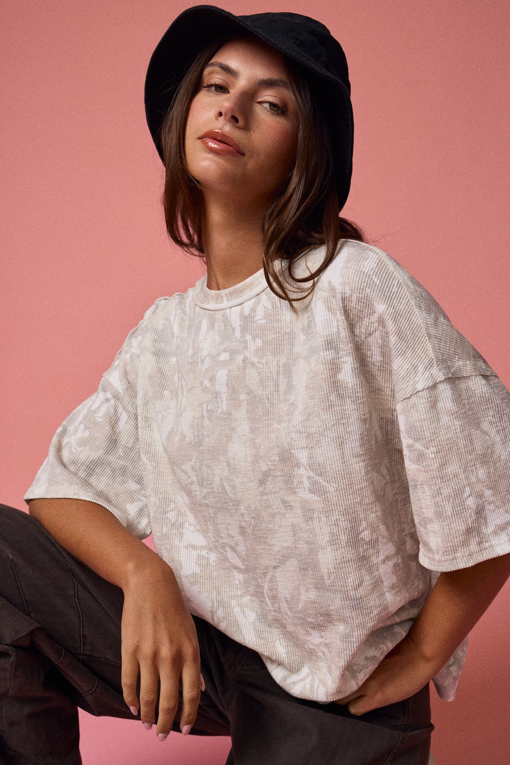 woman wearing a loose-fitting beige floral print top and a black bucket hat against a pink background