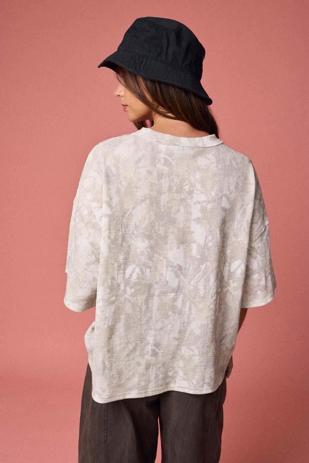 woman wearing an oversized beige tie-dye shirt and a black bucket hat, viewed from the back against a pink background