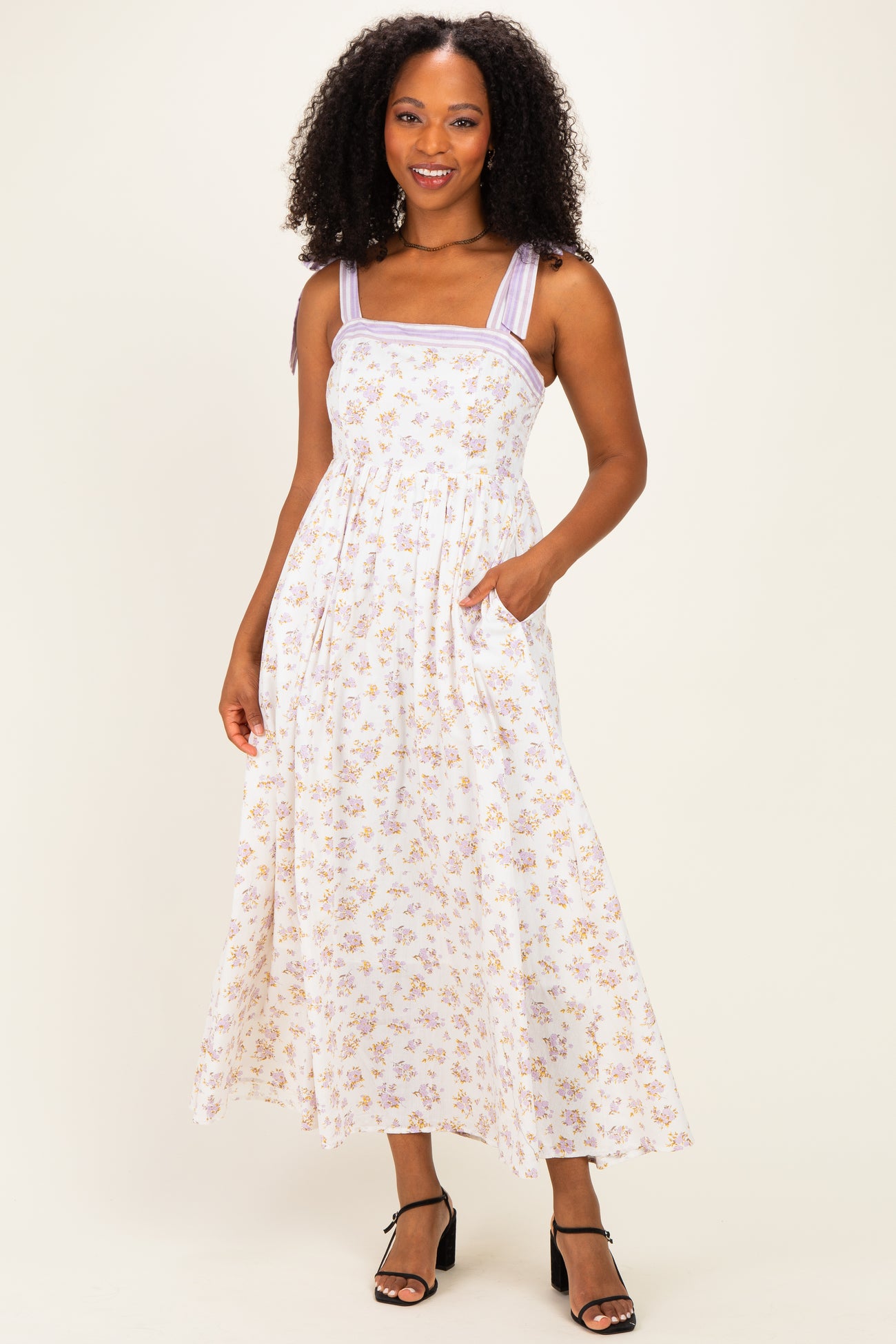 woman wearing a white floral maxi dress with purple straps and pockets, styled with black strappy heels