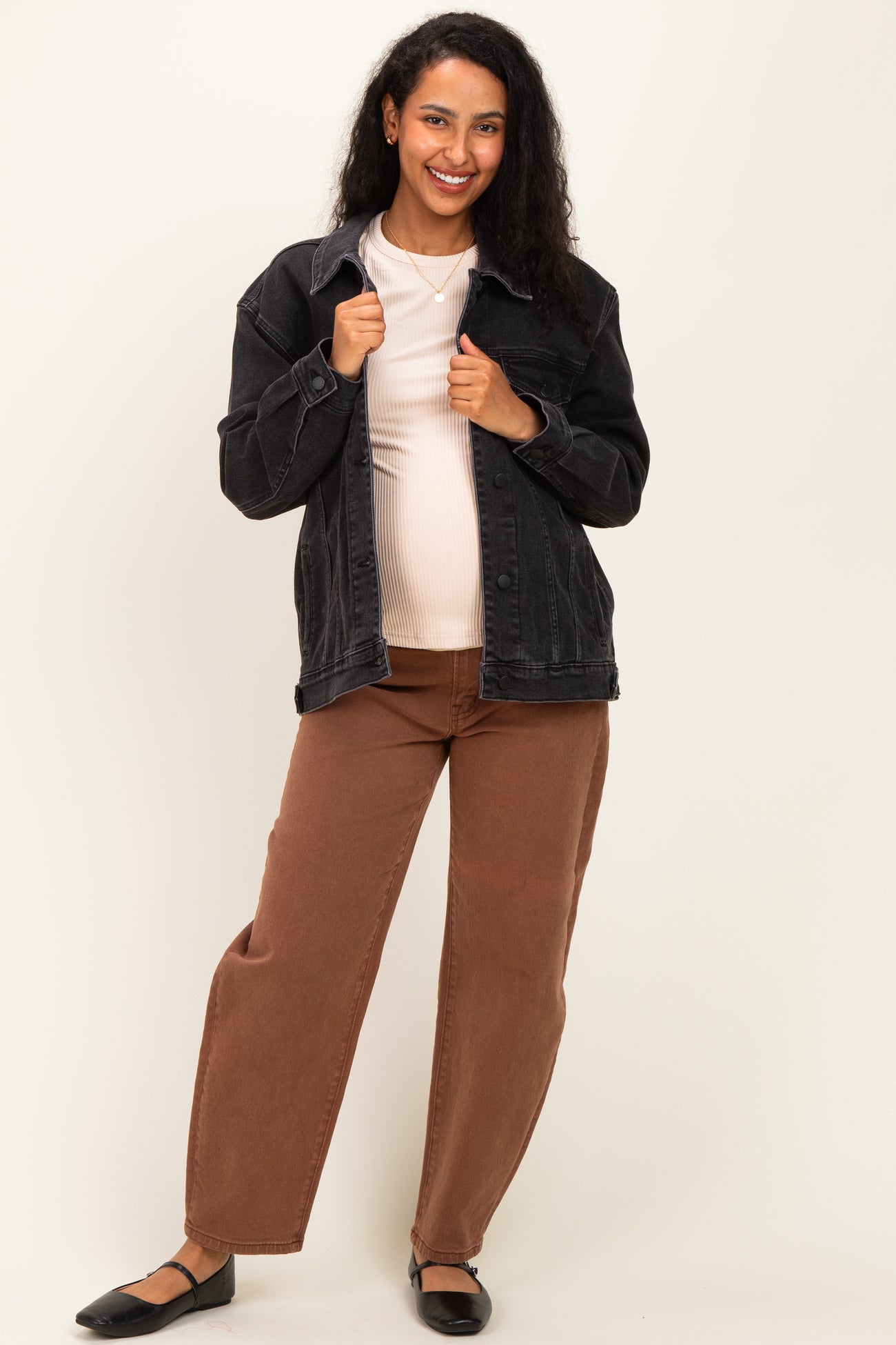 woman wearing a black maternity jacket over a cream top and brown maternity pants, showing her baby bump