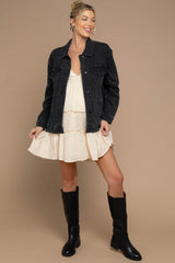 woman wearing a black denim jacket over a cream-colored maternity dress with ruffles and black knee-high boots