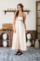 woman wearing a cream maxi dress with thin straps and black detailing, standing in a bright room with a patterned rug