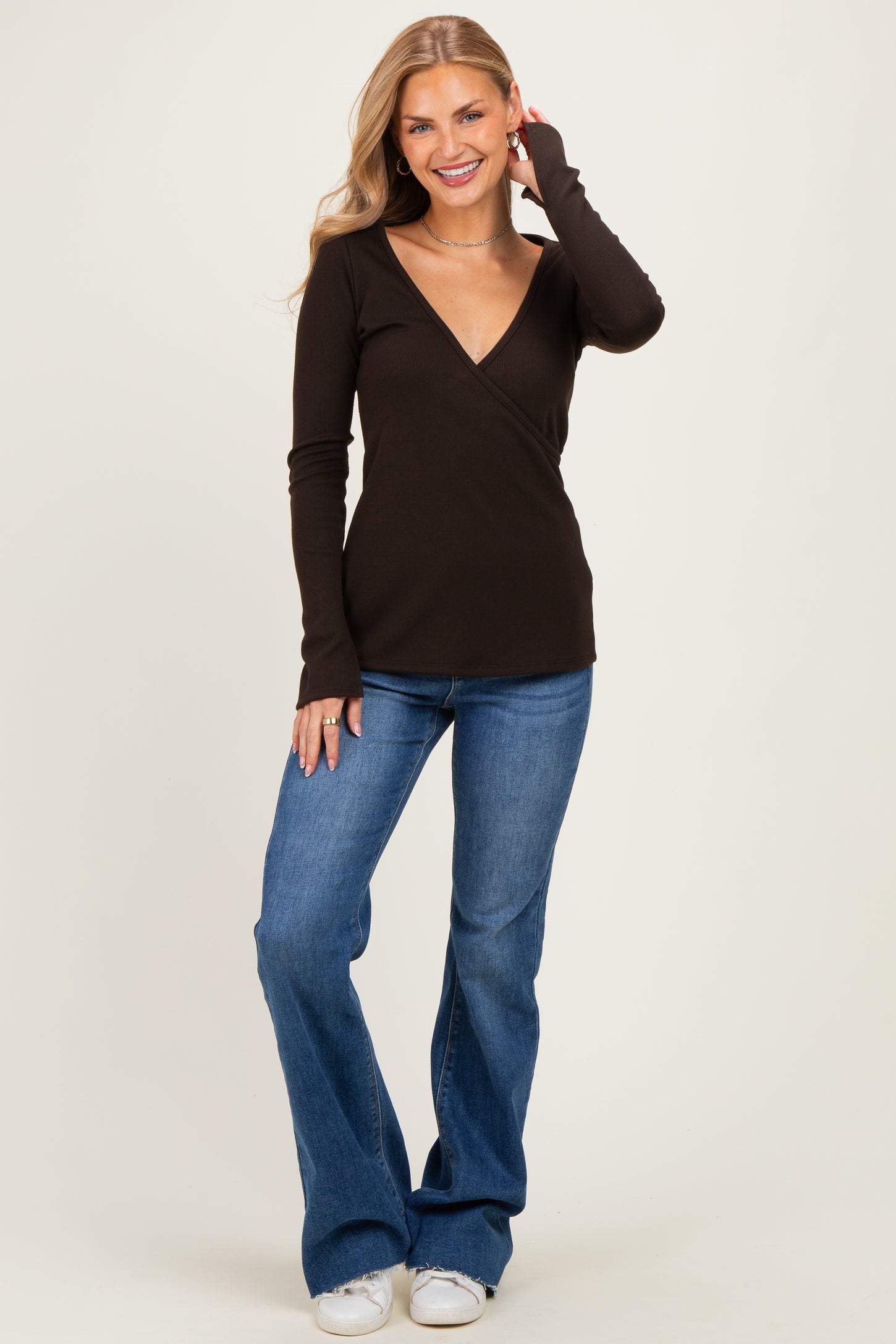 woman wearing a dark brown long sleeve wrap top and flared jeans, smiling and posing against a light background
