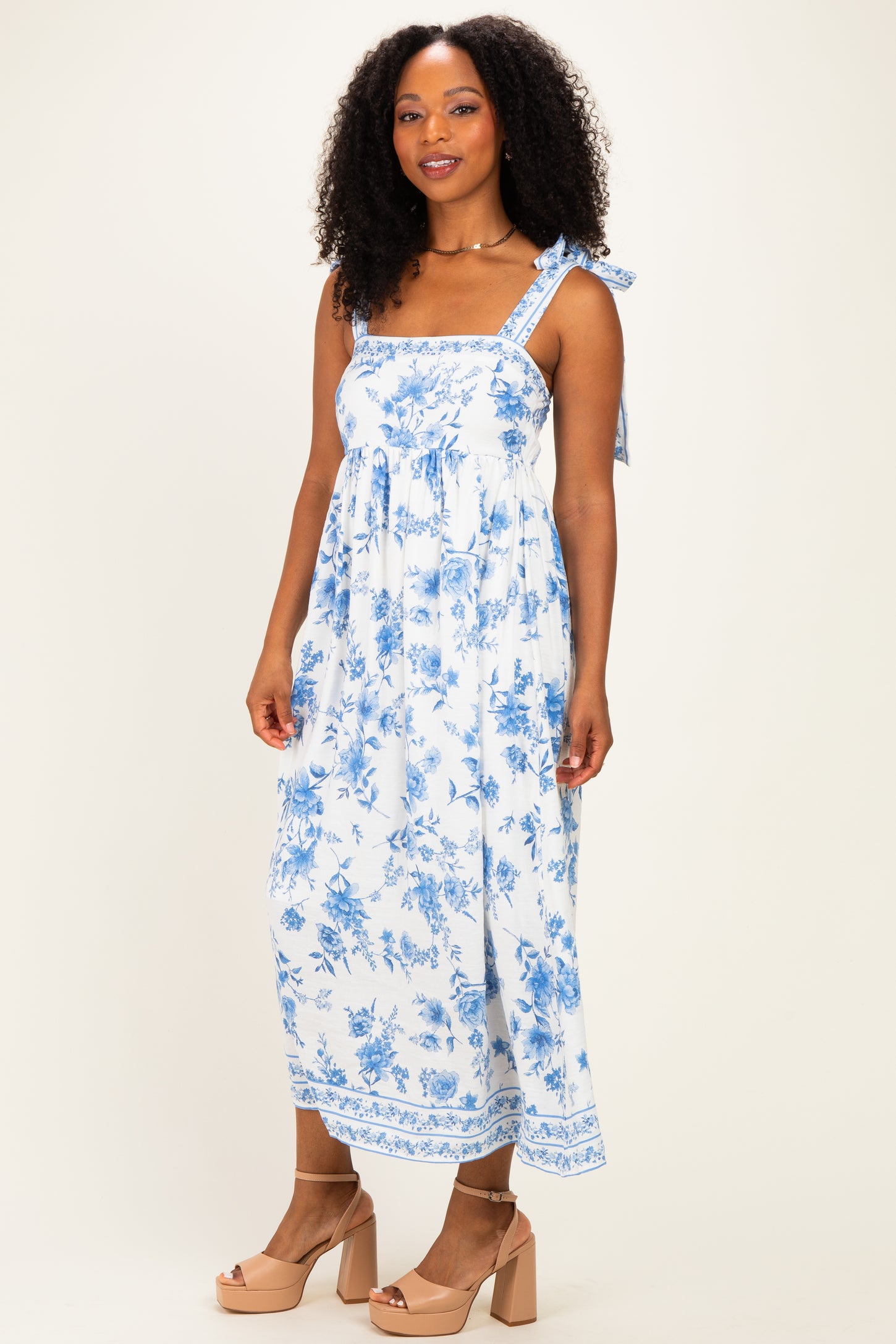 woman wearing a white floral maxi dress with blue patterns and tie straps, standing against a neutral background
