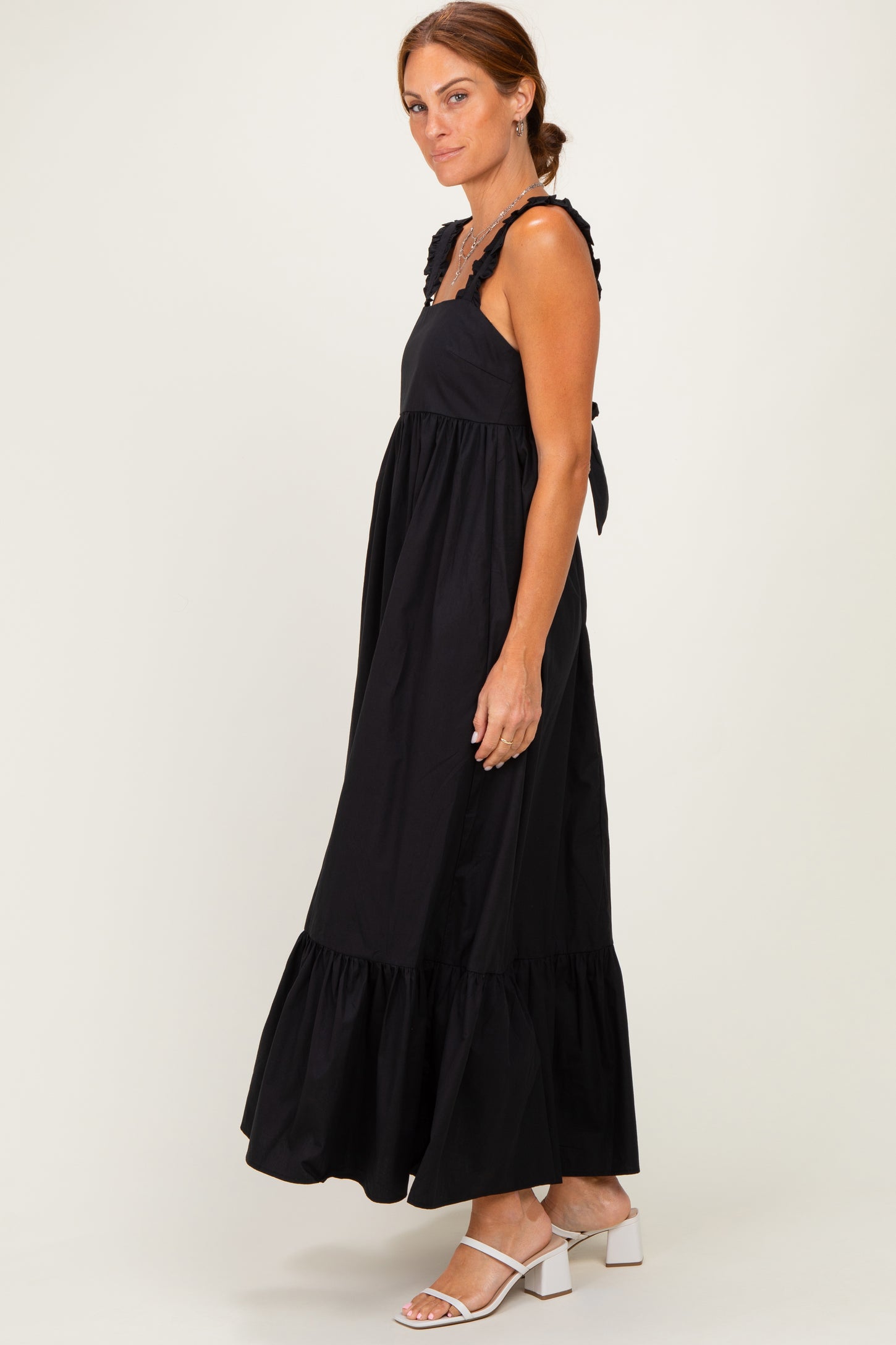 woman wearing a black maxi dress with ruffled straps and a tiered hem, standing sideways with a light background