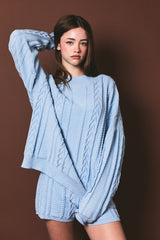 woman wearing a light blue cable knit oversized sweater and matching shorts against a brown background