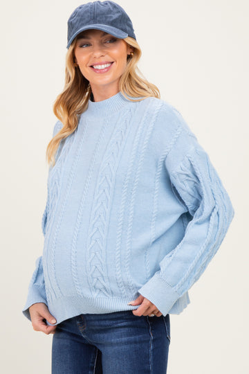 woman wearing a light blue cable knit maternity sweater and a navy cap, showcasing her baby bump