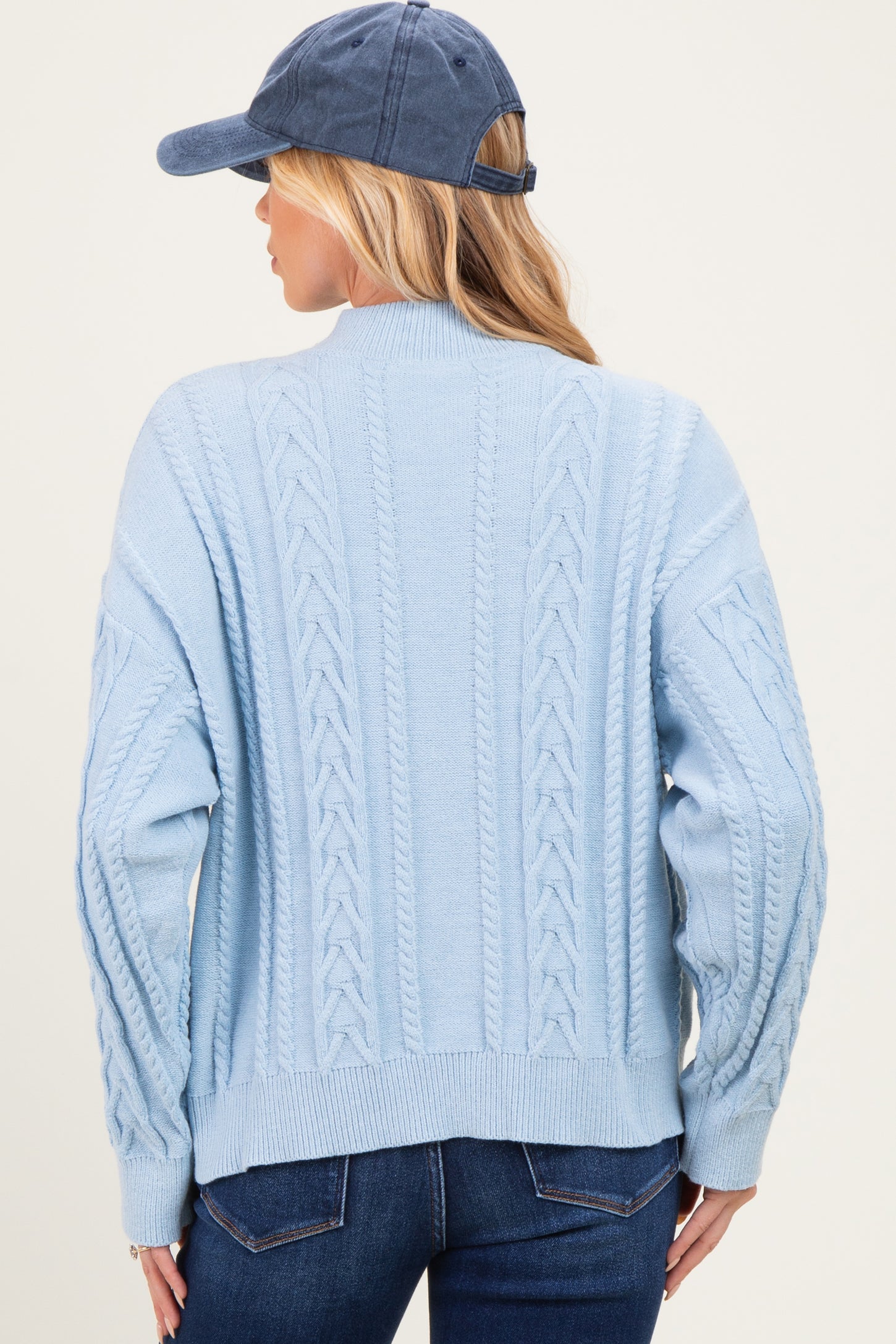 woman wearing a light blue cable knit sweater, view from the back, paired with a navy cap and jeans, maternity wear