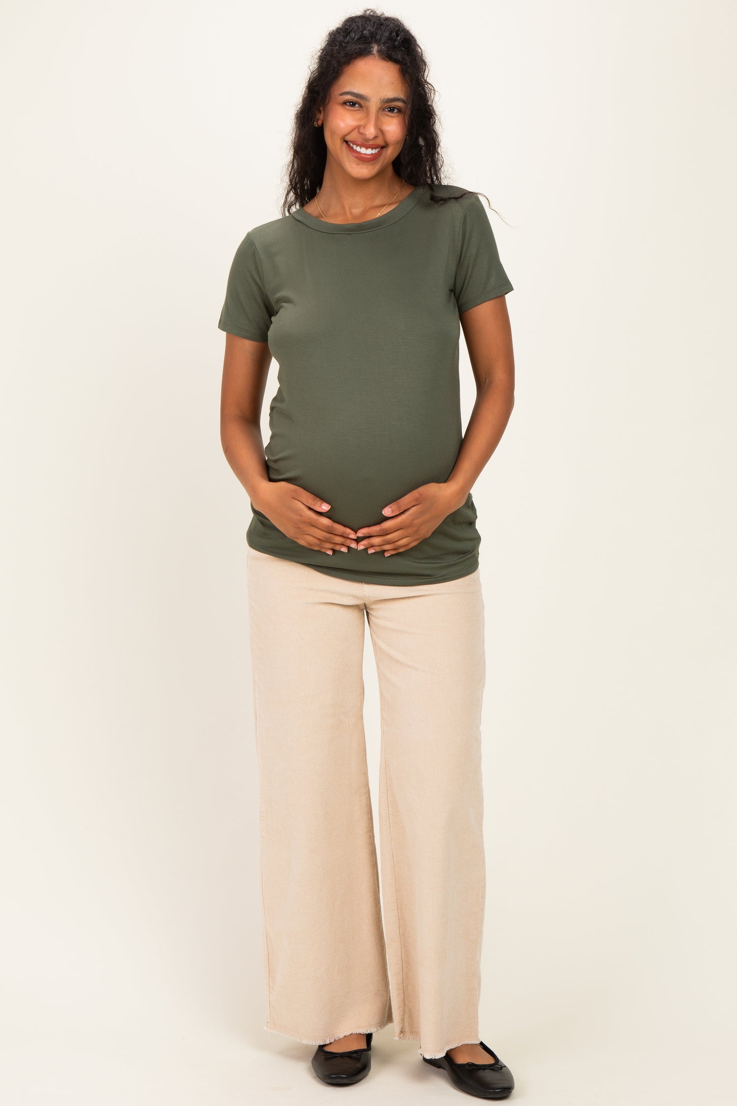 woman wearing an olive green maternity t-shirt and cream wide-leg pants, holding her baby bump