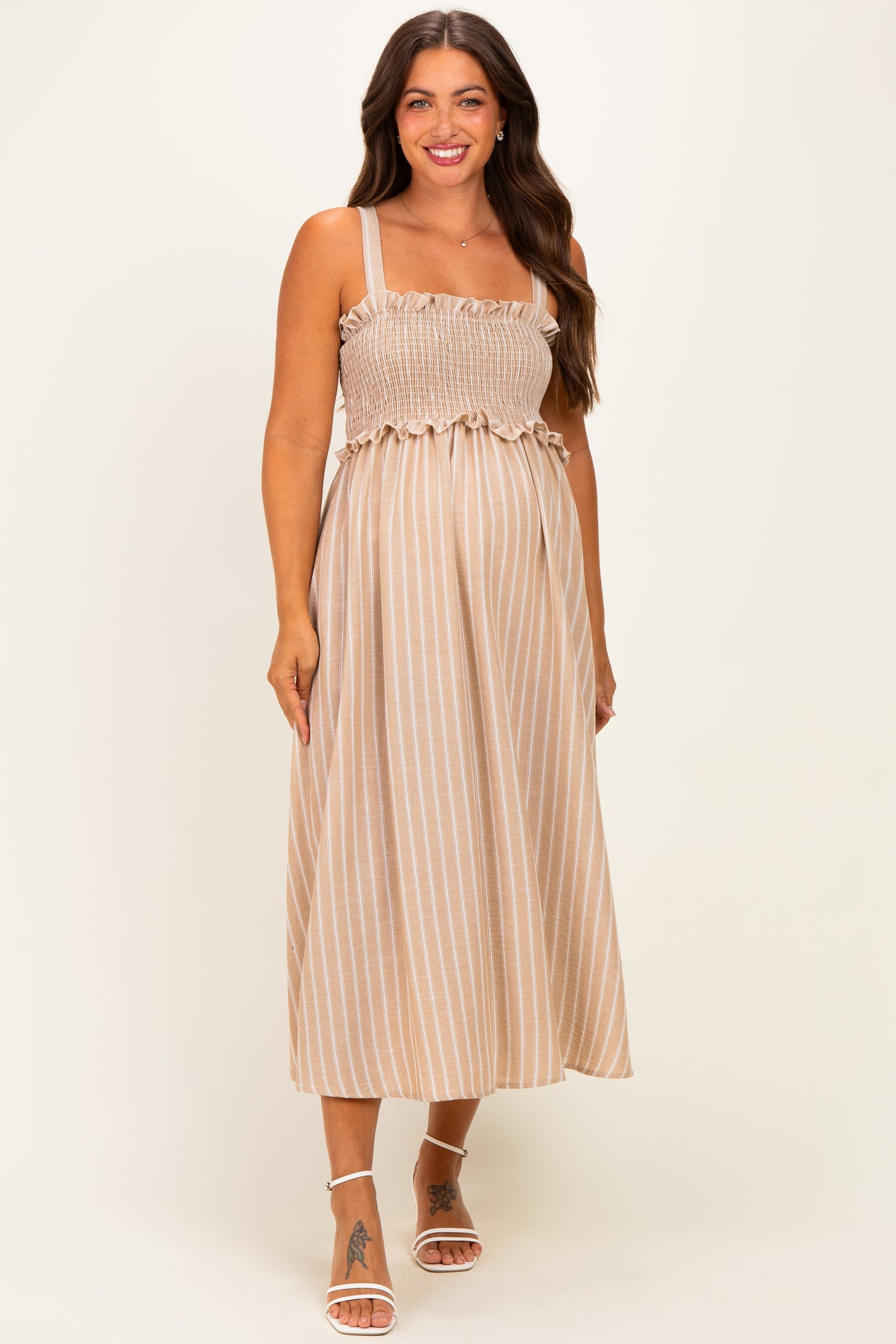 woman wearing a beige striped maternity dress with smocked bodice and adjustable straps, showcasing her baby bump