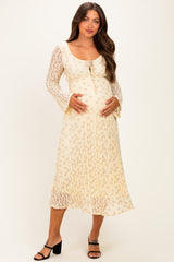 woman wearing a cream floral maternity dress with long lace sleeves and a tie neckline, holding her baby bump