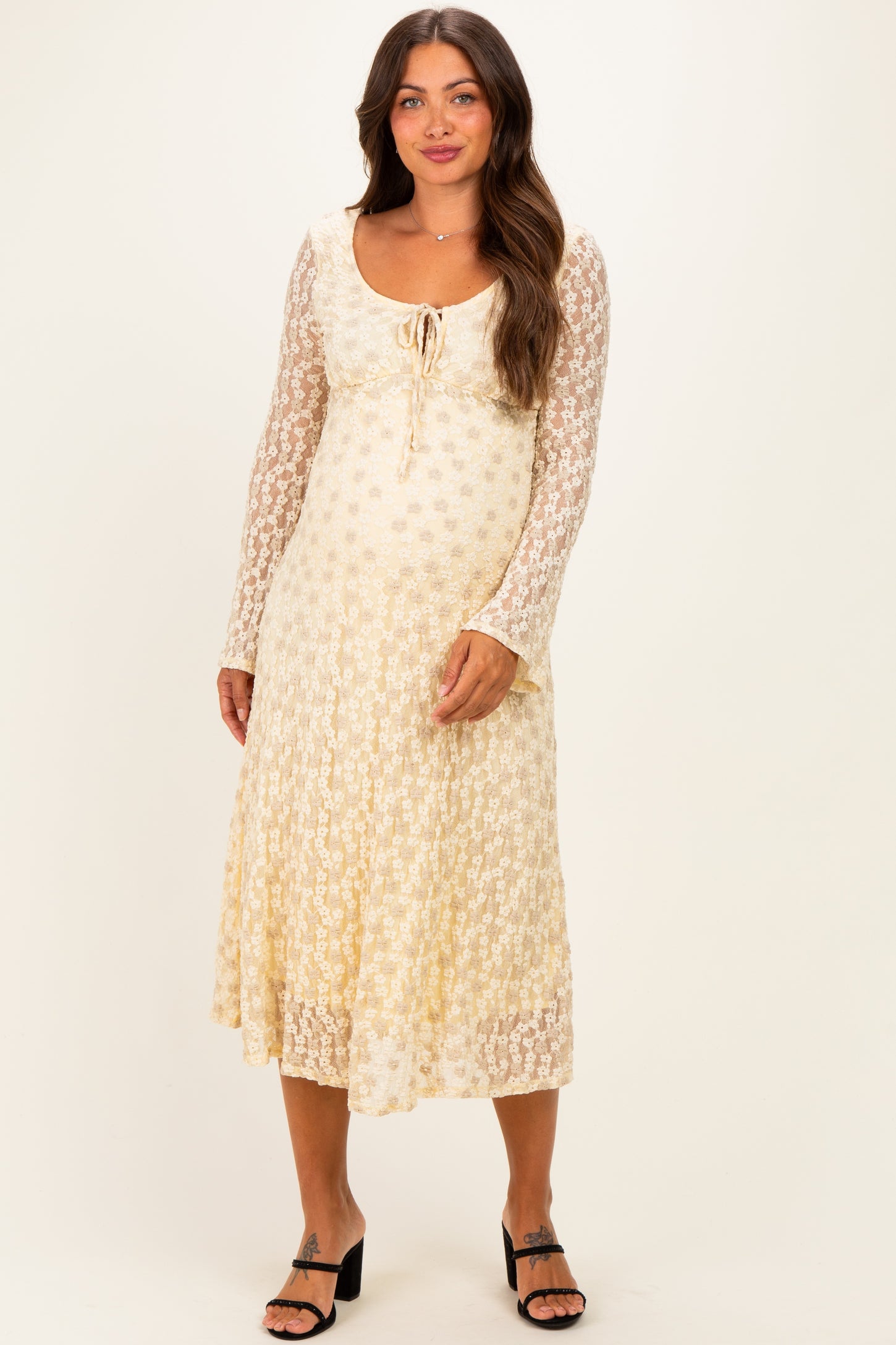 woman wearing a cream floral lace maternity dress with long sleeves and a tie detail at the neckline, showcasing her baby bump