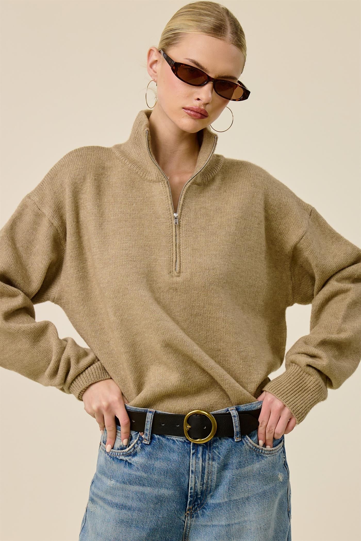 woman wearing a tan quarter zip sweater and high-waisted blue jeans, accessorized with sunglasses and a black belt