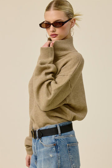 woman wearing a tan turtleneck sweater with oversized sleeves and high-waisted distressed jeans, accessorized with sunglasses