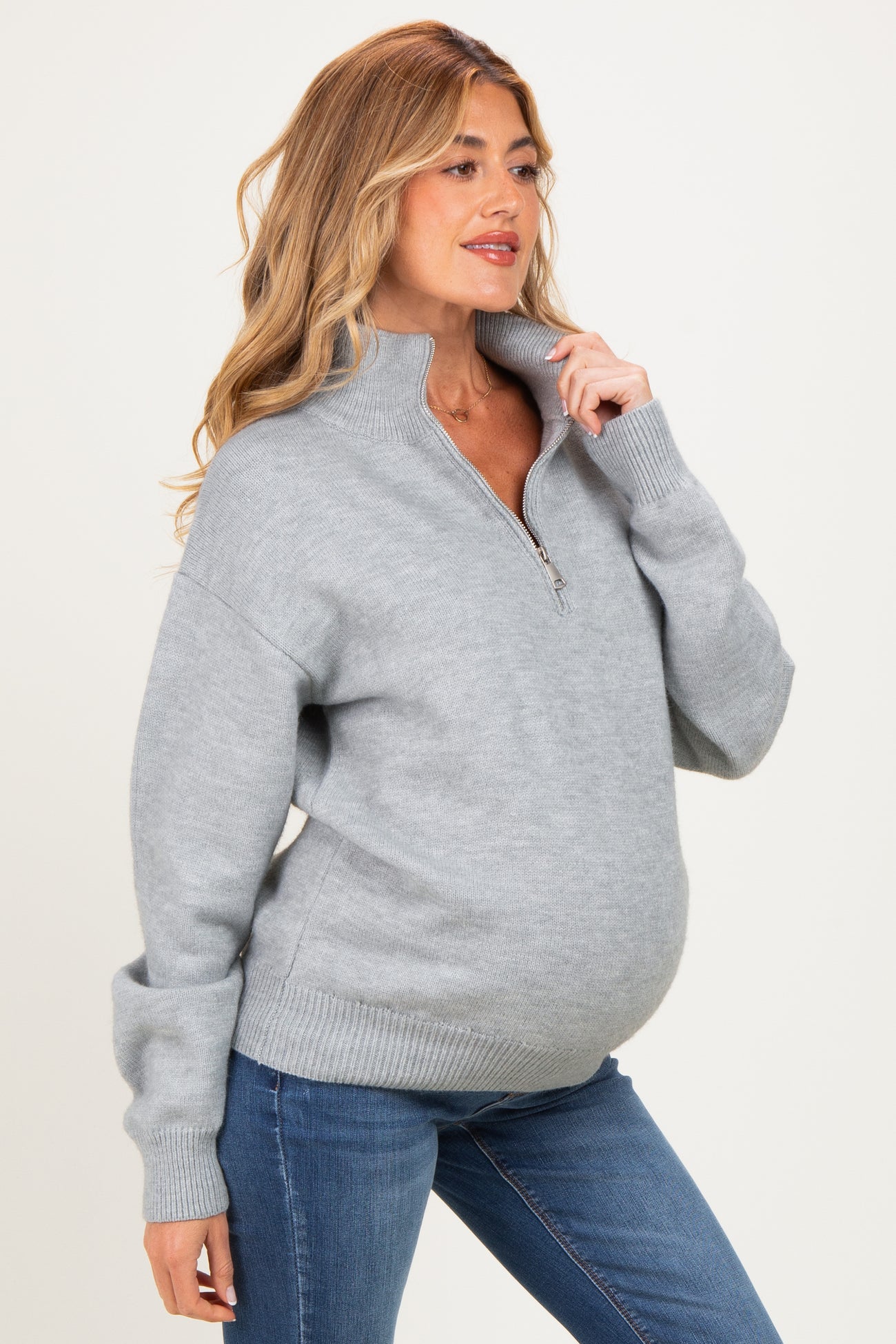 woman wearing a light grey zip-up maternity sweater with a relaxed fit and visible baby bump, paired with blue jeans