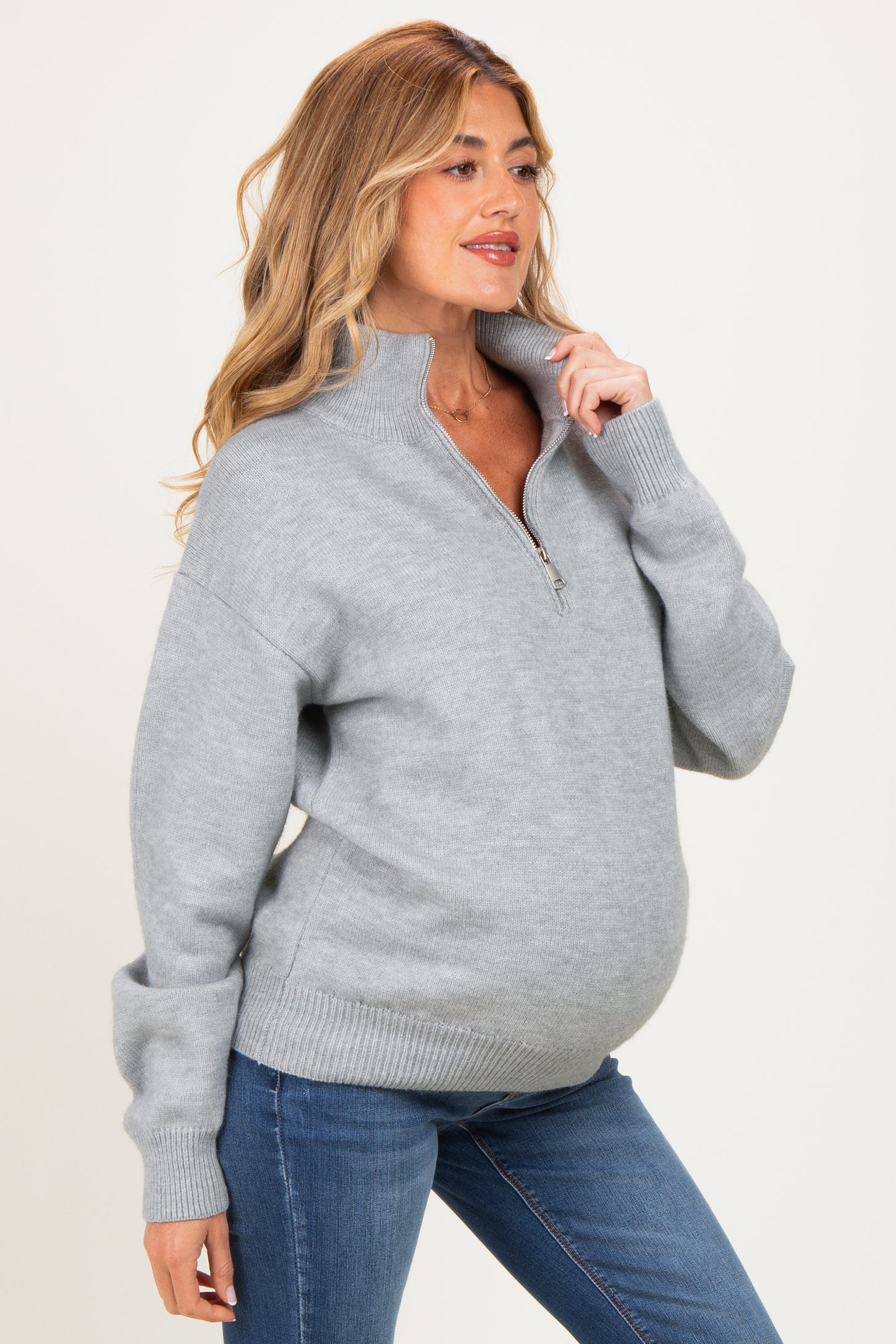 woman wearing a light grey zip-up maternity sweater with a relaxed fit and visible baby bump, paired with blue jeans