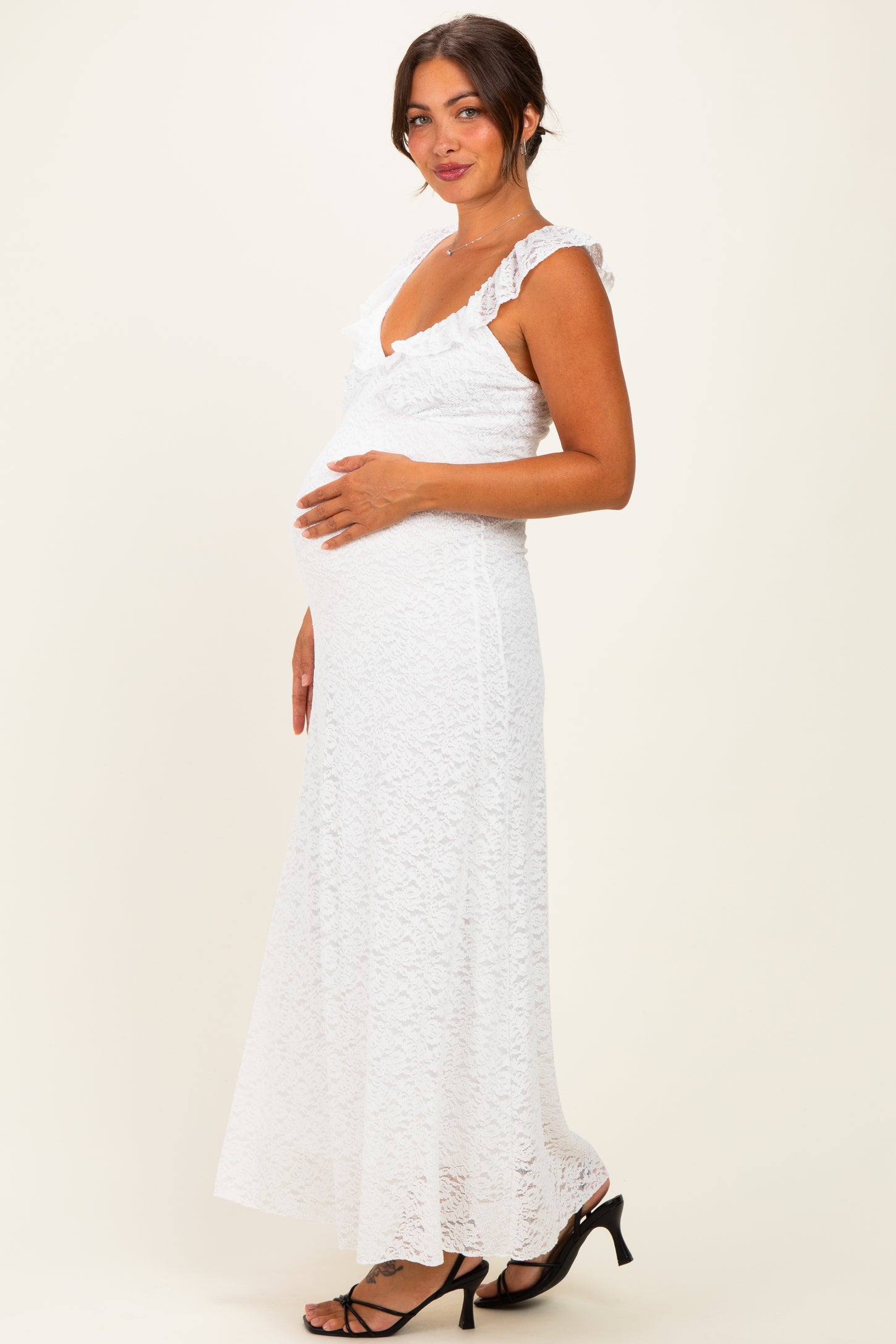 woman wearing a white lace maternity maxi dress, gently holding her baby bump and smiling, with ruffled straps