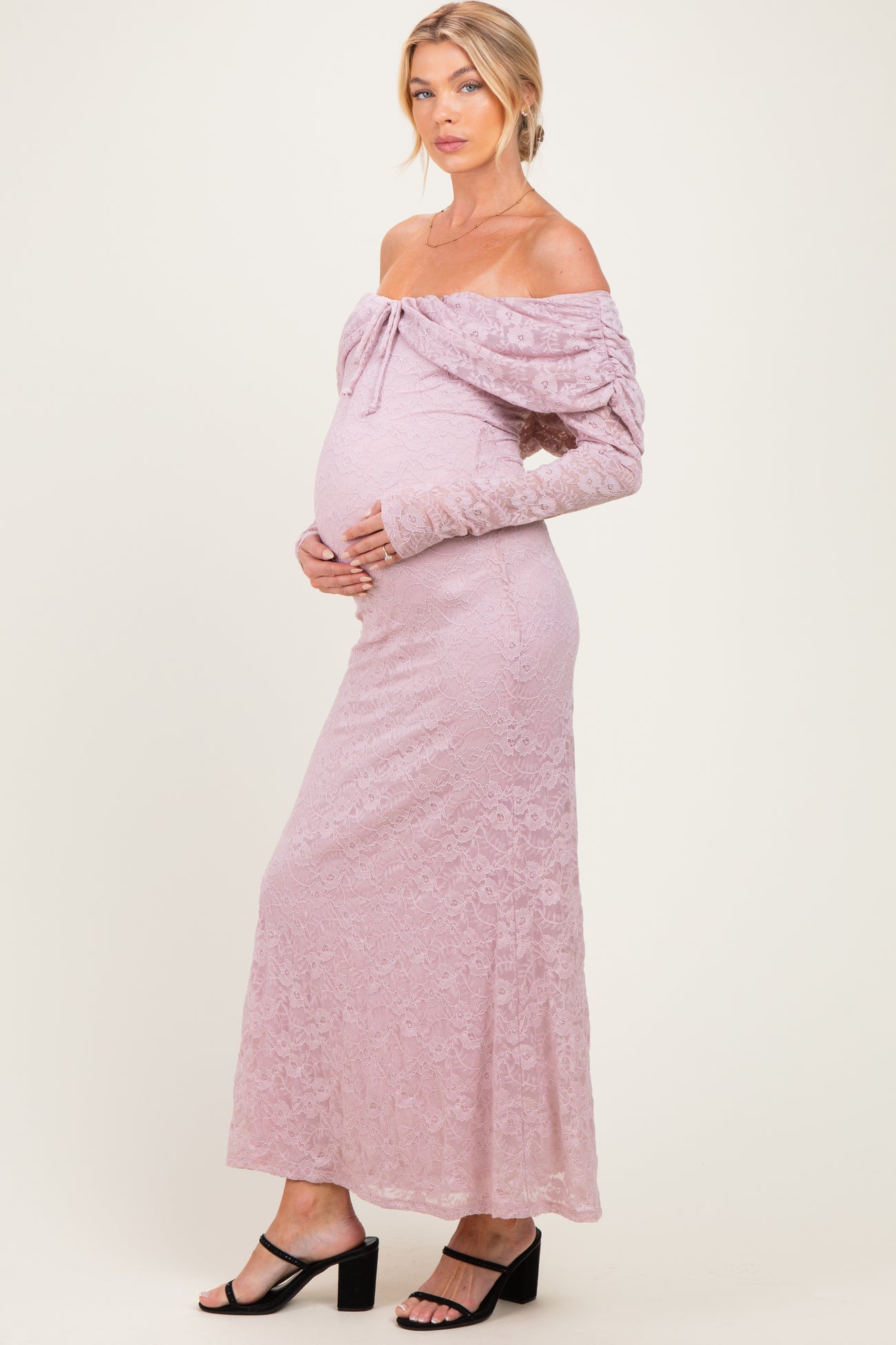 woman wearing a light pink off-the-shoulder lace maternity dress, gently cradling her baby bump