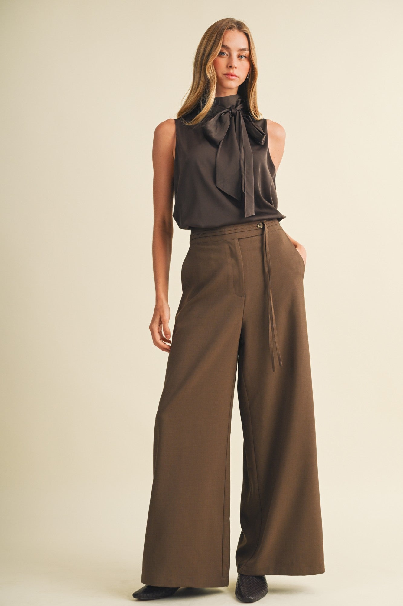 woman wearing a dark brown sleeveless blouse with a bow tie and wide-leg brown trousers