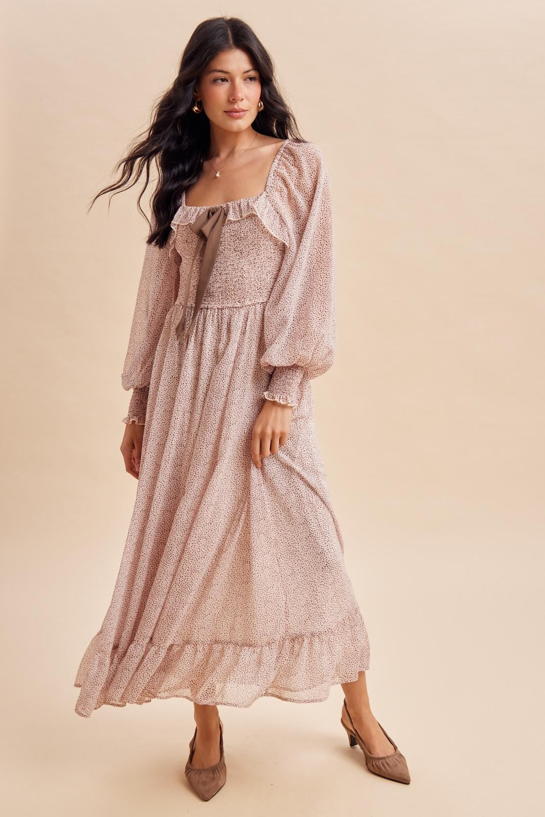 woman wearing a blush long sleeve maxi dress with ruffle details and a bow at the neckline, styled with brown heels