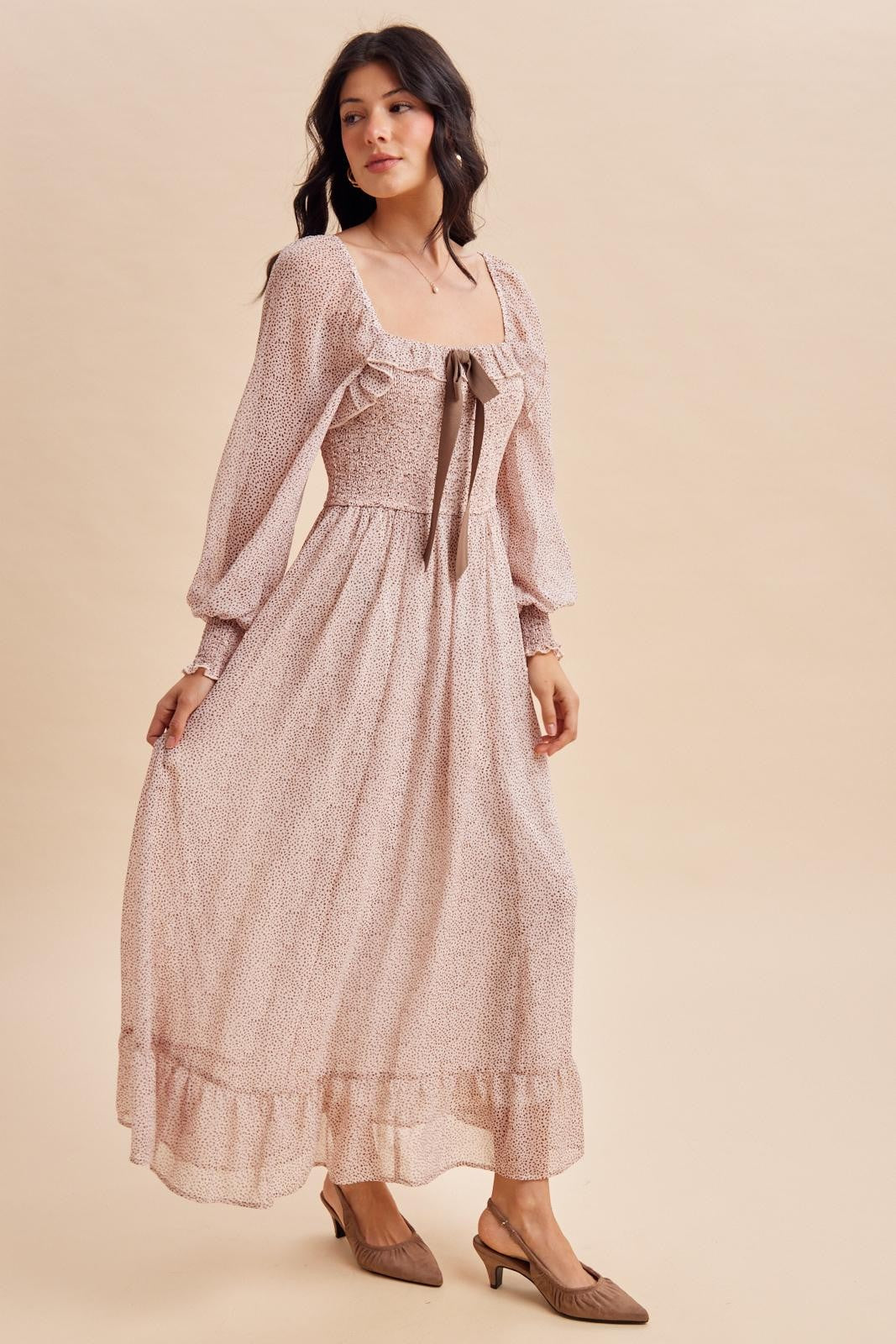 woman wearing a blush floral print maxi dress with puff sleeves and a bow detail at the neckline, paired with beige mules