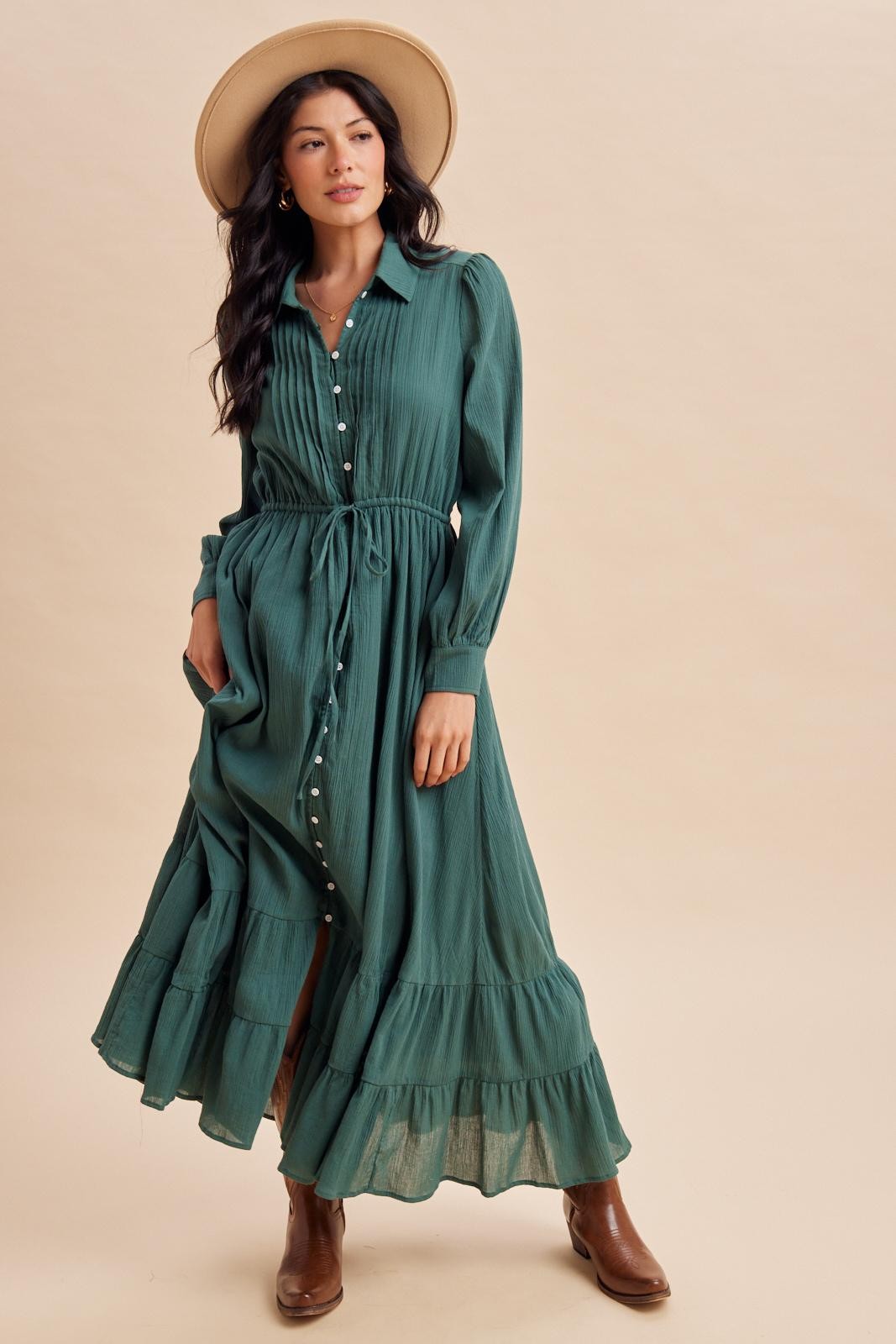 woman wearing a green long sleeve button-up maxi dress with a tie waist and tiered skirt, paired with a wide-brimmed hat and brown boots