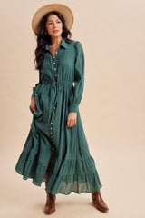 woman wearing a green long sleeve button-up maxi dress with a tie waist and tiered skirt, paired with a wide-brimmed hat and brown boots