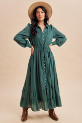 woman wearing a green long sleeve button-up maxi dress with a ruffled hem and a tie waist, accessorized with a hat and brown boots