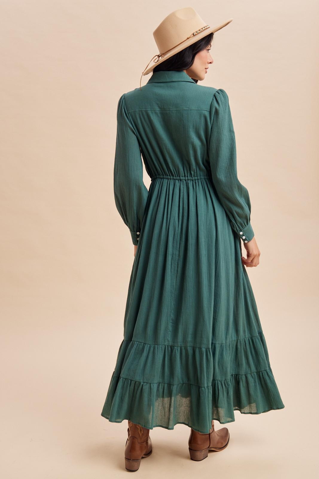 woman wearing a long green dress with puff sleeves and a tiered skirt, accessorized with a beige hat and brown boots