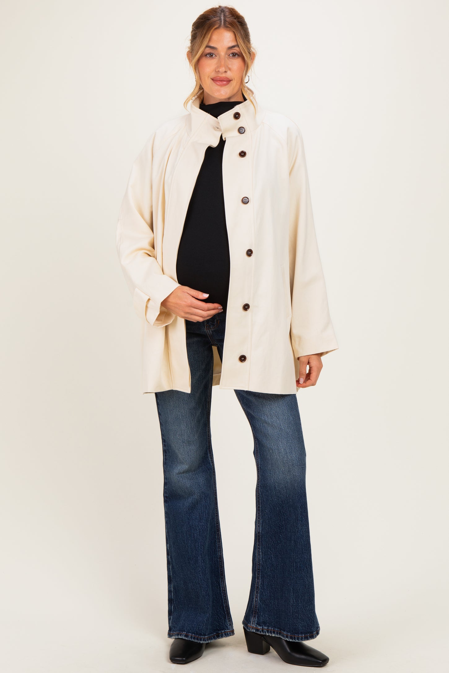 woman wearing a cream maternity jacket over a black top and blue flared jeans, holding her baby bump
