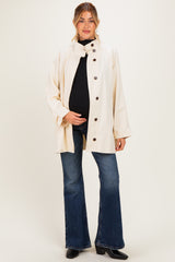 woman wearing a cream maternity jacket over a black top and blue flared jeans, holding her baby bump