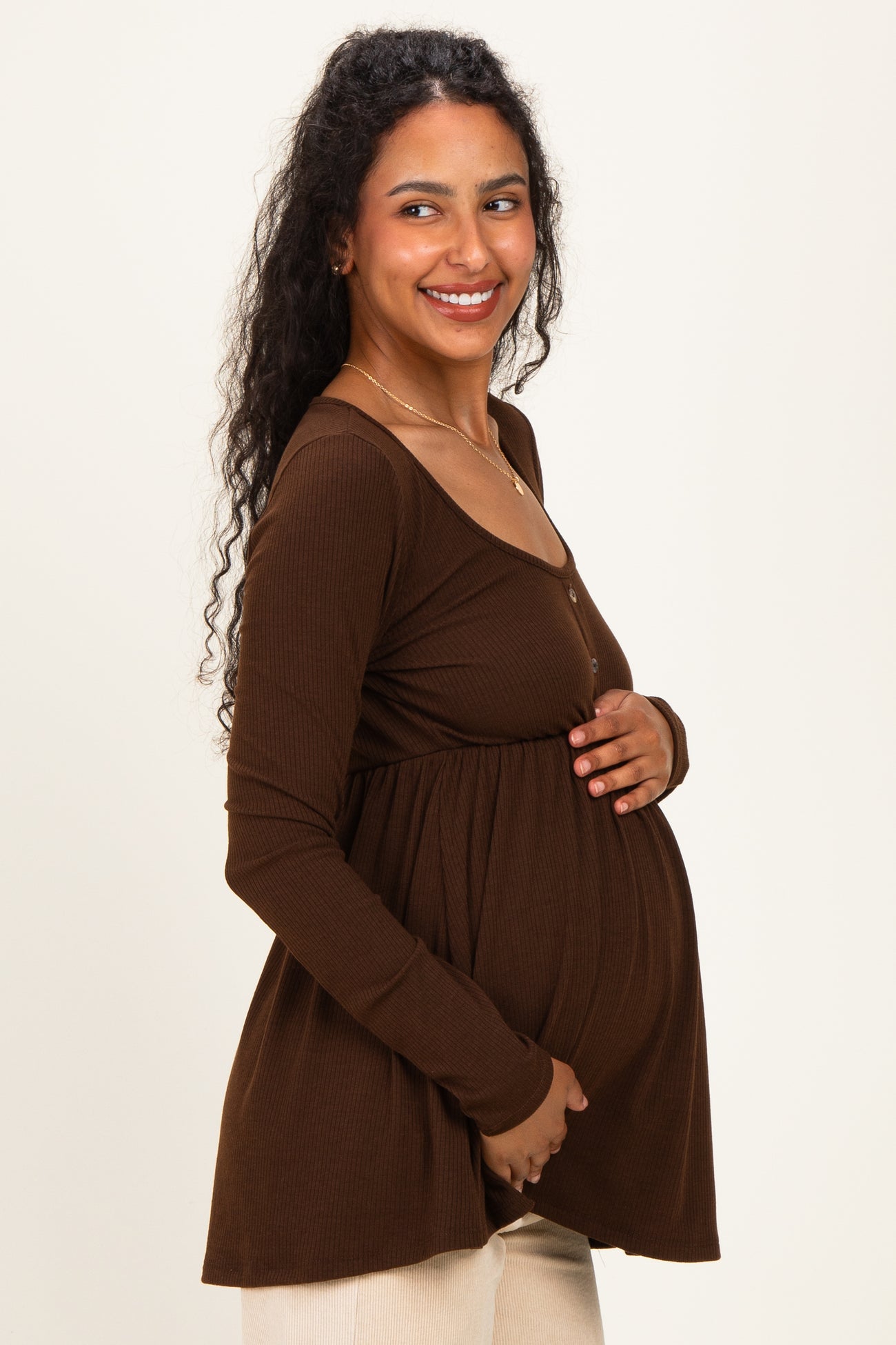 woman wearing a brown long sleeve maternity top, gently holding her baby bump while smiling