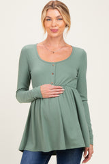 woman wearing a sage green long sleeve maternity top with a scoop neckline and button details, holding her baby bump