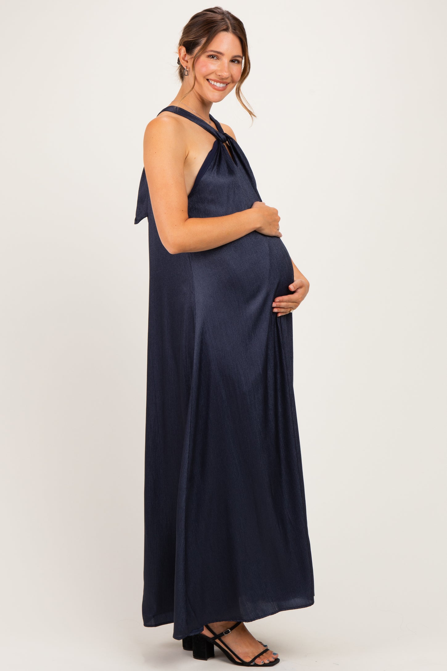 woman wearing a navy blue sleeveless maternity maxi dress with a tie back and holding her baby bump