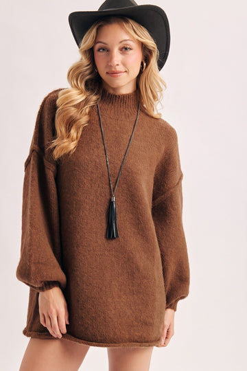 woman wearing a brown oversized sweater dress with long sleeves and a high neckline, accessorized with a black hat